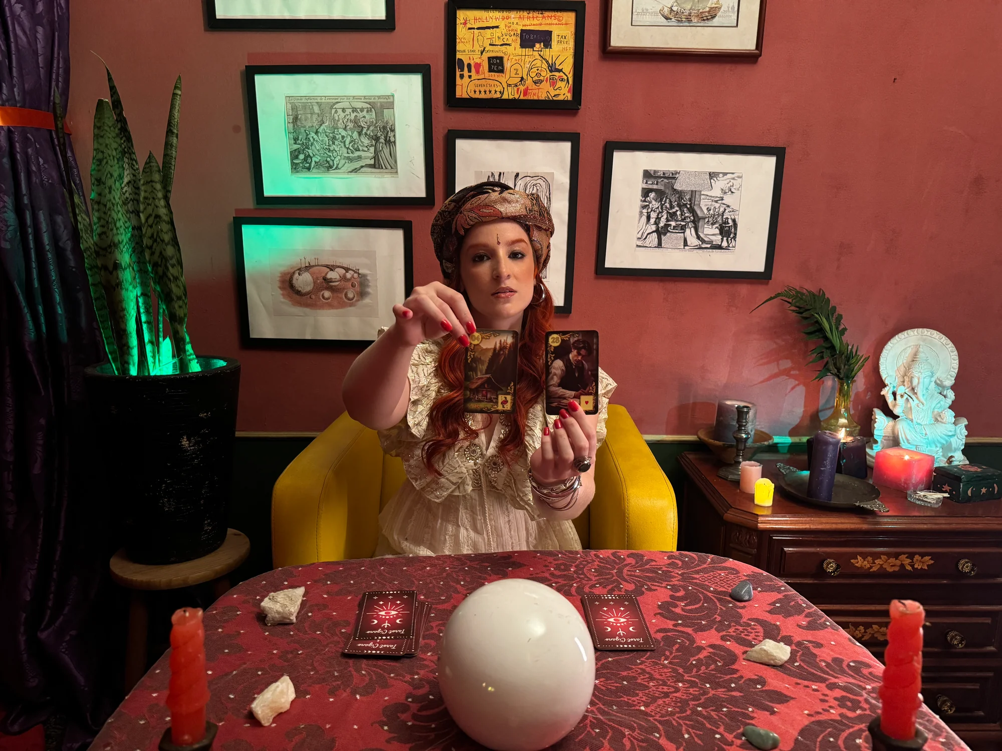 Orange Girl de turbante segurando duas cartas de tarô diante de uma mesa com bola de cristal e velas